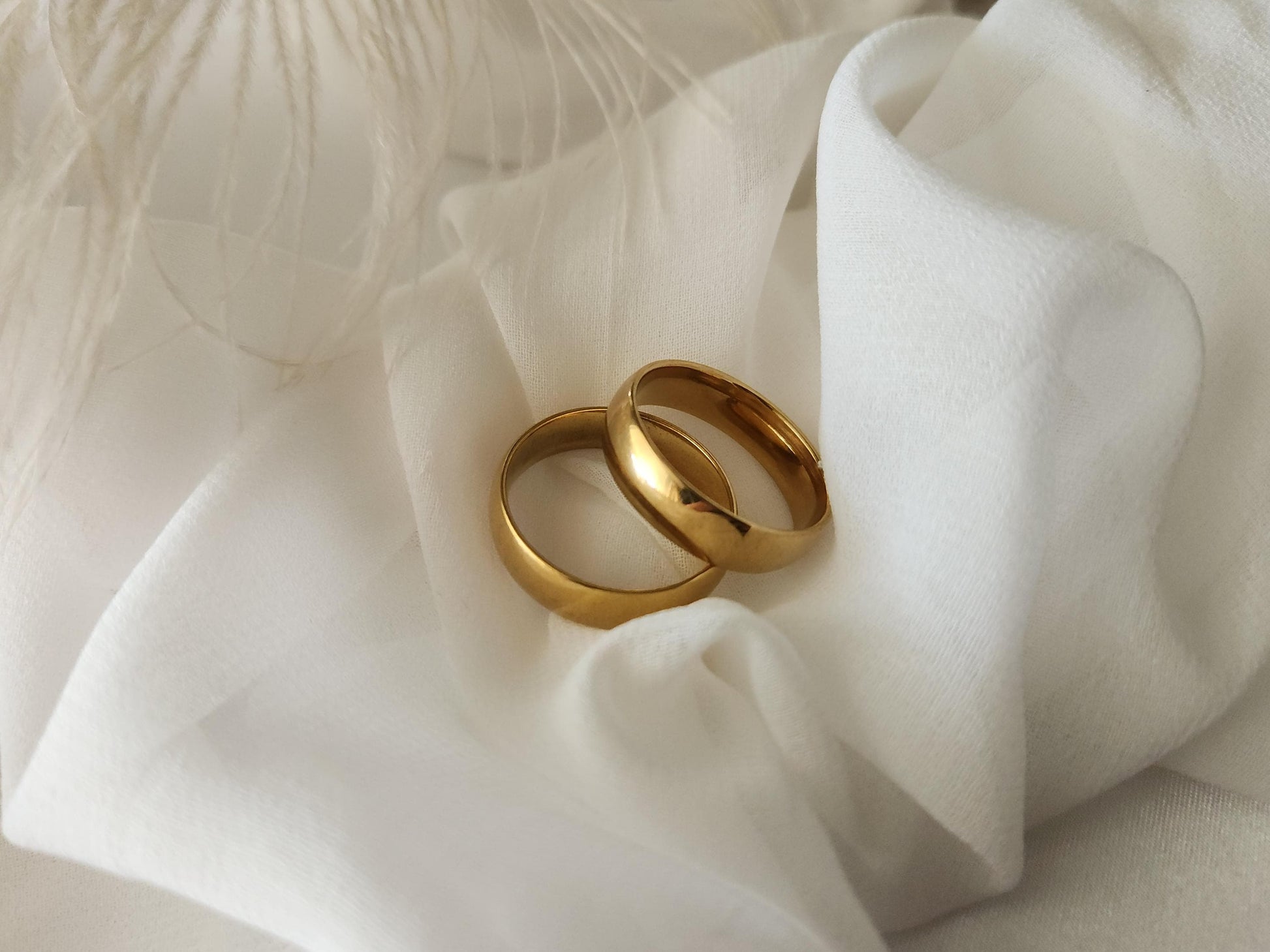 May include: Two gold-colored wedding bands rest on a white fabric background. The bands are simple, smooth, and have a high polish finish.  They are positioned slightly overlapping, suggesting a pair. The bands appear to be made of metal. The fabric is soft and has subtle folds and creases. The overall aesthetic is minimalist and elegant, suitable for showcasing wedding jewelry.
